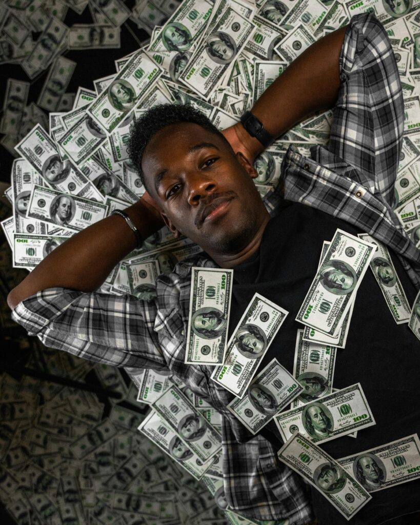 Man in plaid shirt lying on a bed of US dollar bills, showcasing wealth and abundance.
